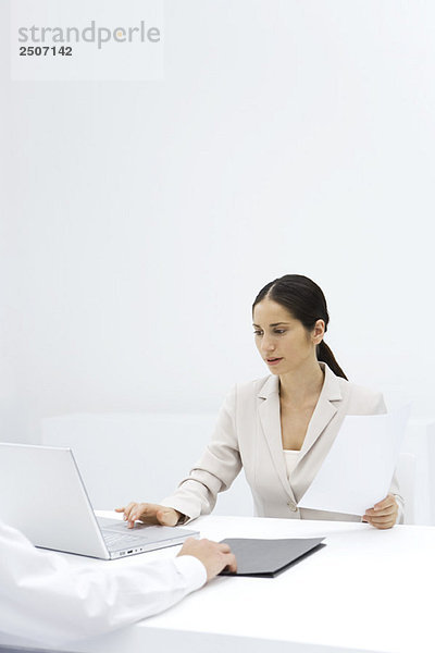 Frau  die einen Laptop benutzt  während sie ein Dokument hält  beschnittene Ansicht des Arms  der den Ordner über den Schreibtisch schiebt.
