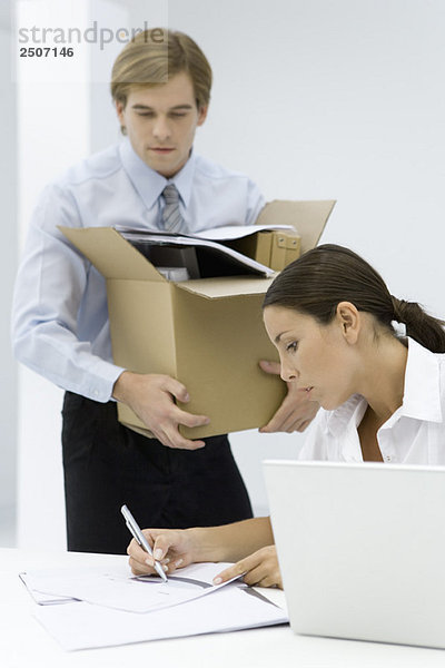 Frau schreibt auf Dokument am Schreibtisch  Mann trägt Pappkarton voll mit Bürobedarf