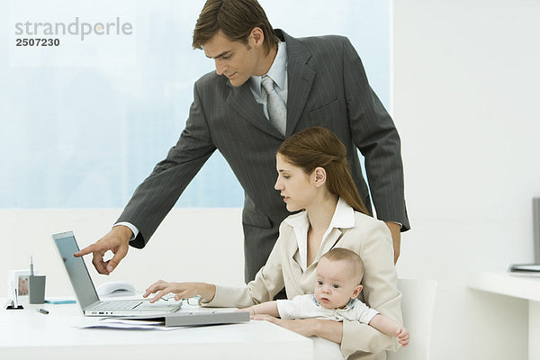 Professionelle Frau im Büro  Baby auf dem Schoß haltend  Laptop mit männlichem Kollegen betrachtend
