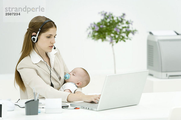 Geschäftsfrau im Büro  hält schlafendes Baby  benutzt Headset und Laptop Computer