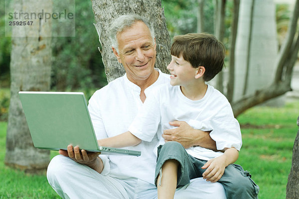 Großvater hält Enkel auf Schoß  Junge schaut auf Laptop-Computer  beide lächelnd