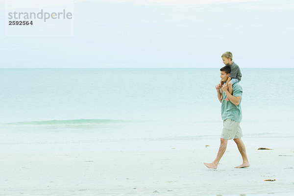 Ein Mann  der am leeren Strand spazieren geht und seinen Sohn auf den Schultern trägt.