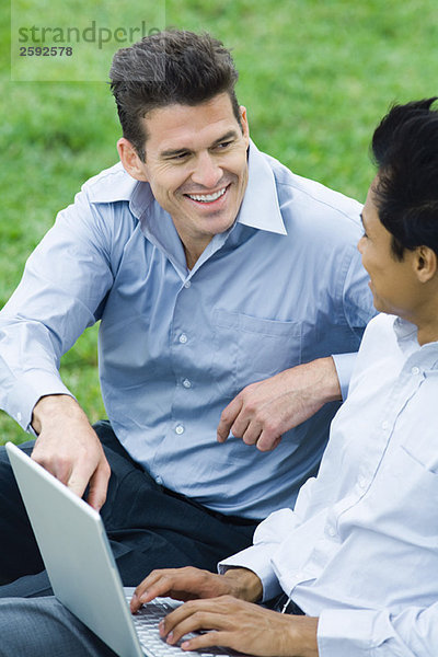 Zwei Männer sitzen im Park mit Laptop und sehen sich lächelnd an.