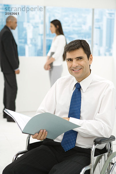 Professioneller Mann im Rollstuhl mit Dokumenten  lächelnd in die Kamera  Kollegen im Hintergrund