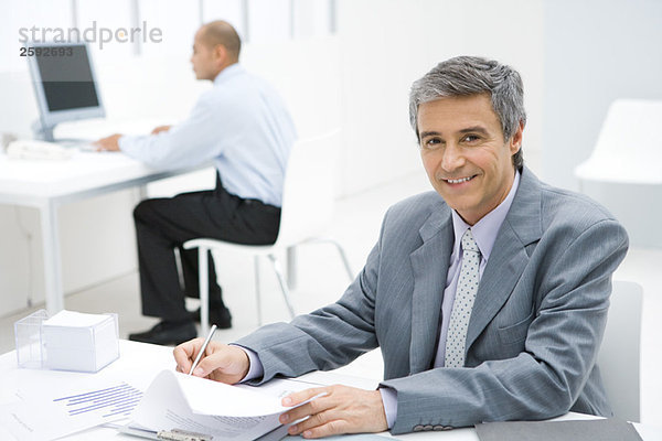Geschäftsmann am Schreibtisch  lächelnd vor der Kamera  Kollege im Hintergrund
