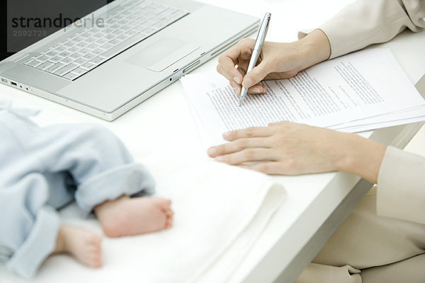 Frau redigiert Dokument am Schreibtisch  Kleinkind liegt in der Nähe  Schnittansicht
