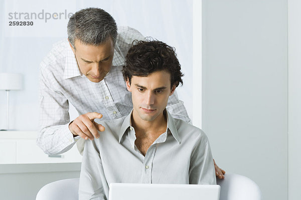 Reifer Mann  der sich über die Schulter des jungen Mannes lehnt  beide mit Blick auf den Laptop.
