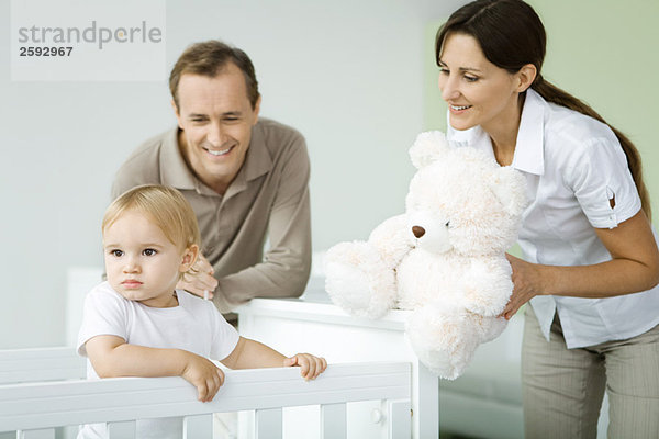 Eltern stehen neben Kleinkind im Kinderbett  Mutter hält Teddybär  Kleinkind schaut weg