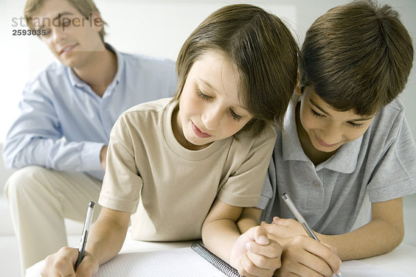 Zwei Jungen schreiben zusammen in ein Notizbuch  Vater sieht im Hintergrund zu.