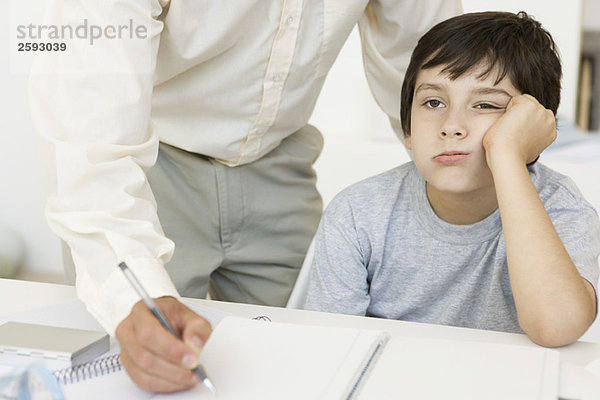 Vater hilft dem Sohn bei den Hausaufgaben  Junge hält den Kopf und schaut weg  Blick in den Ausschnitt