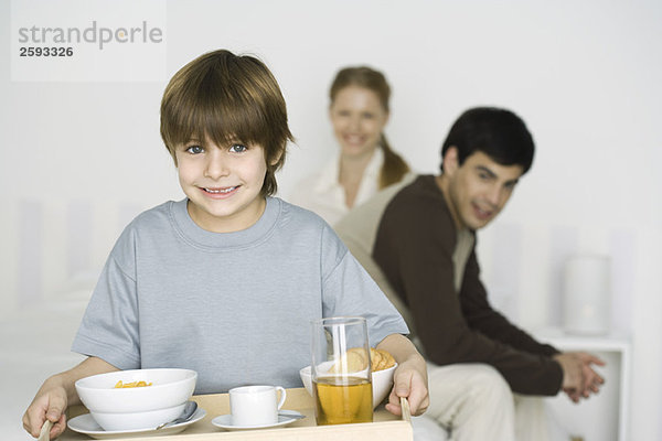 Kleiner Junge mit Frühstück auf Tablett  Eltern sitzen im Hintergrund auf dem Bett.