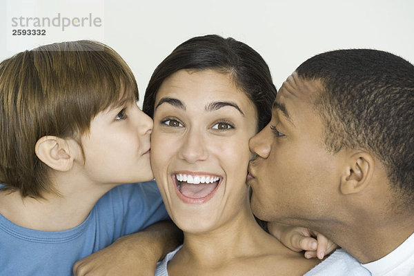 Frau lächelt vor der Kamera als Mann und Sohn ihre Wangen küssen