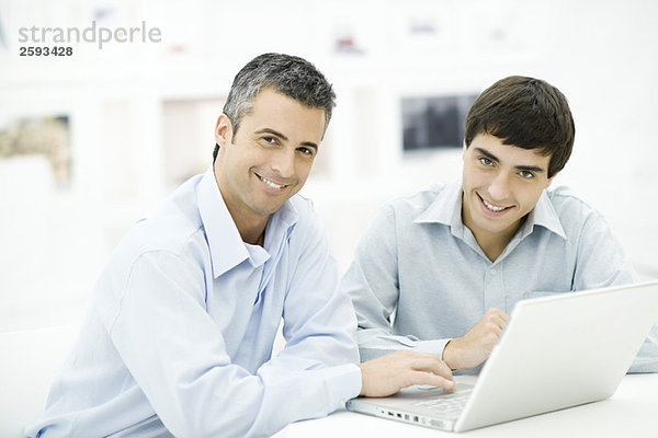 Zwei Männer  die zusammen einen Laptop benutzen  beide lächeln in die Kamera.