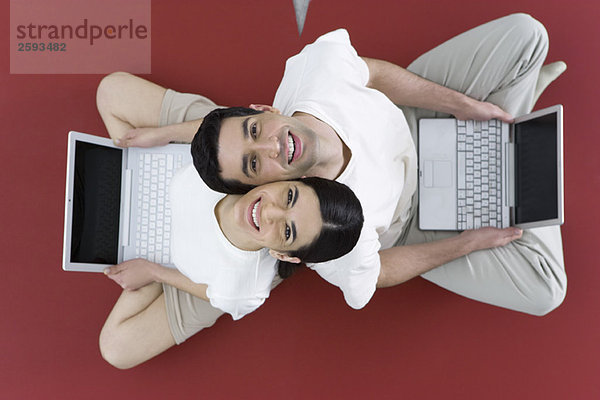 Paar sitzt Rücken an Rücken auf dem Boden  hält Laptop-Computer und lächelt vor der Kamera.