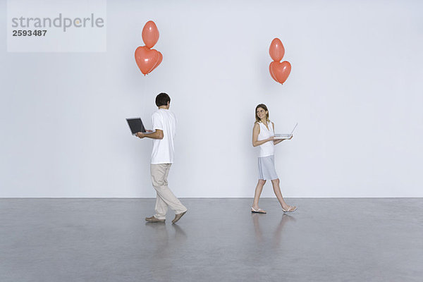 Mann und Frau tragen Laptop-Computer und Herzballons und lächeln sich gegenseitig über die Schultern.