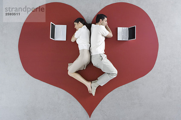 Mann und Frau liegen Rücken an Rücken auf großem Herzen  beide mit Blick auf Laptops.