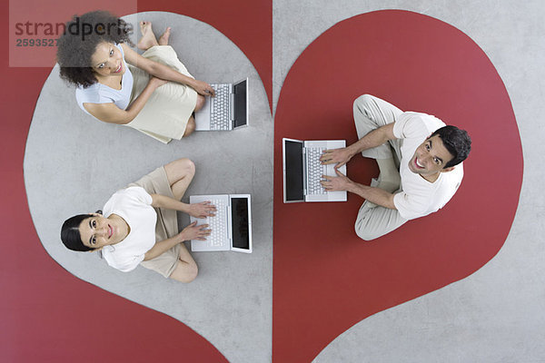 Zwei Frauen und ein Mann sitzen auf einem großen Herzen  benutzen Laptops und lächeln vor der Kamera.