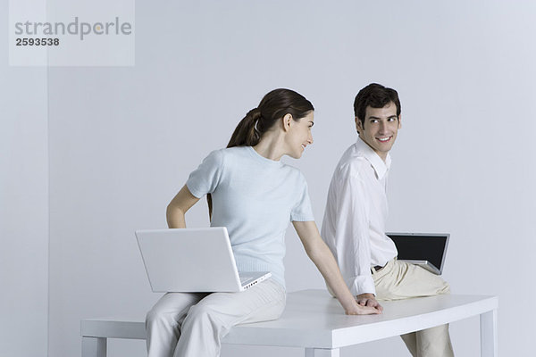 Ein Paar sitzt Rücken an Rücken mit Laptops auf dem Schoß  hält sich an den Händen und lächelt sich an.
