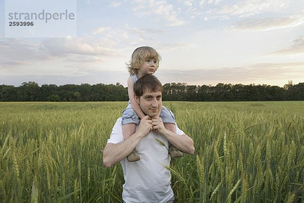 Ein Mann  der ein Kind auf seinen Schultern durch ein Weizenfeld trägt.