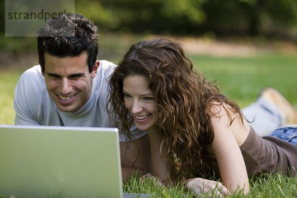 Ein junges Paar  das auf der Vorderseite liegt und auf einen Laptop schaut und lacht.