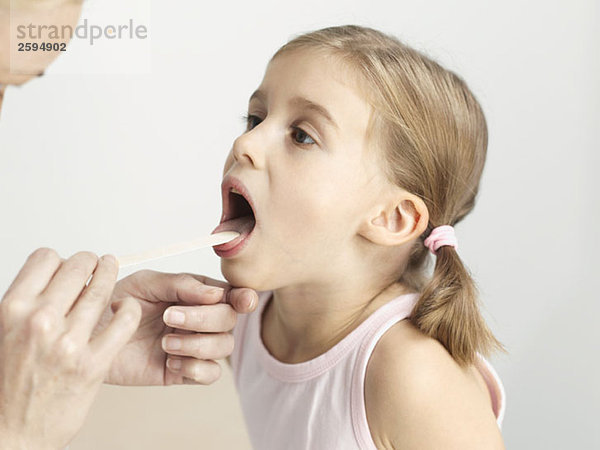 Ein junges Mädchen  das seine Kehle mit einem Zungenspatel untersuchen lässt.