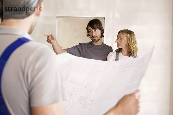 Bauarbeiter betrachtet Bauplan  junges Paar im Hintergrund