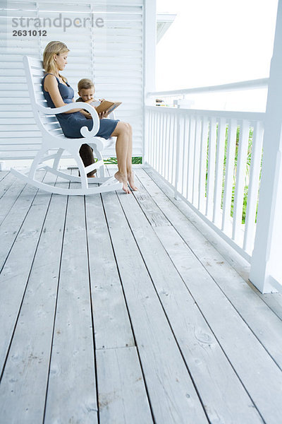 Frau sitzt im Schaukelstuhl auf der Veranda und liest dem kleinen Jungen  der an ihrer Seite steht  die Geschichte vor.
