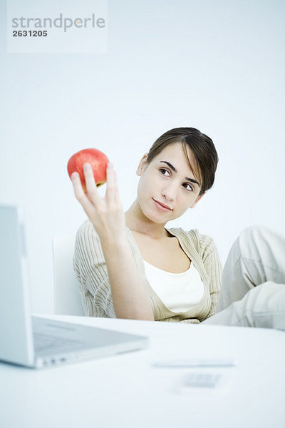 Junge Frau sitzt am Schreibtisch und schaut auf den Apfel in der Hand.