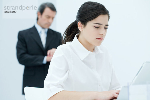 Junge Frau beim Tippen auf dem Laptop  Chef überprüft die Zeit im Hintergrund