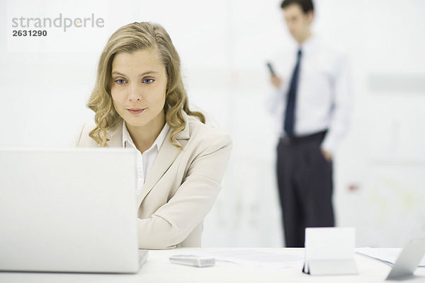 Professionelle Frau mit Laptop am Schreibtisch  Mann steht im Hintergrund