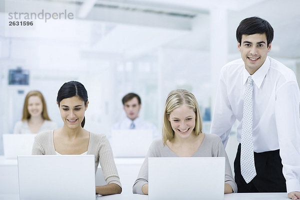 Vorgesetzter steht neben Mitarbeitern  die an Laptops arbeiten und lächelt in die Kamera.