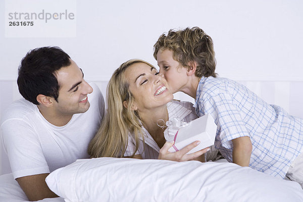 Frau im Bett liegend mit Mann und Sohn  Geschenk haltend  Junge küssend ihre Wange