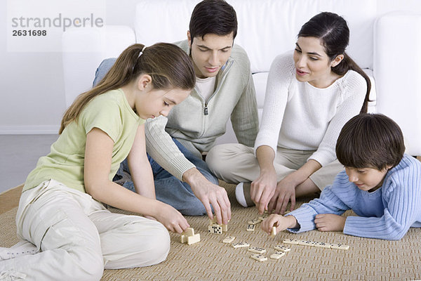 Eltern und zwei Kinder sitzen auf dem Boden und spielen zusammen Dominosteine.