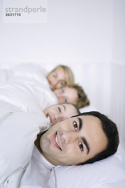 Mann liegt mit seiner Familie im Bett und lächelt in die Kamera.