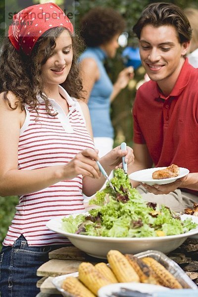 Junge Frau verteilt Blattsalat auf Teller beim Grillfest