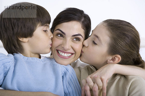 Porträt einer lächelnden Mutter  die von Tochter und Sohn auf jede Wange geküsst wird.