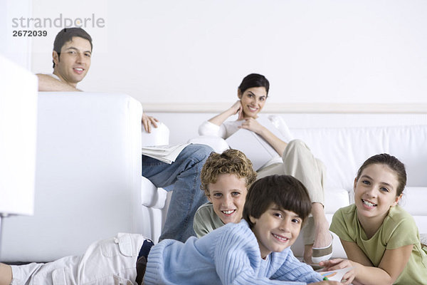 Familie zusammen im Wohnzimmer  Eltern sitzen auf dem Sofa  Kinder liegen auf dem Boden  lächeln in der Kamera