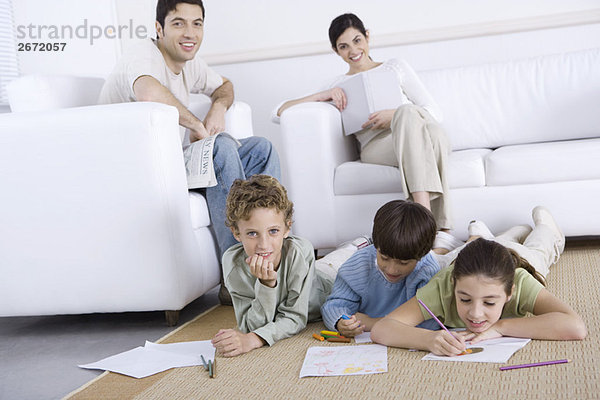 Drei Kinder malen auf dem Boden im Wohnzimmer  Eltern entspannen im Hintergrund