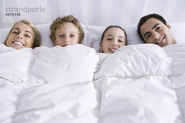 Familie liegt zusammen im Bett unter der Bettdecke und lächelt in die Kamera.
