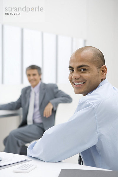 Geschäftsmann am Schreibtisch sitzend  über die Schulter lächelnd vor der Kamera  Kollege im Hintergrund