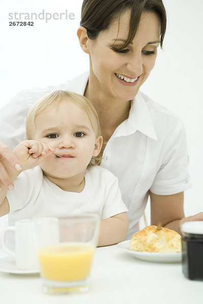 Professionelle Frau sitzt am Frühstückstisch  Kleinkind sitzt auf dem Schoß  Löffel im Mund