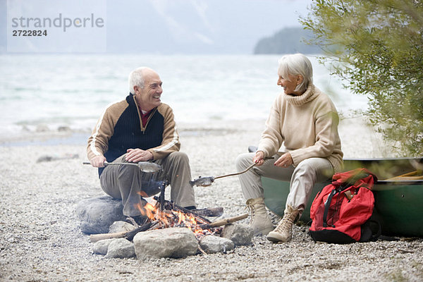 Germany  Bavaria  Walchensee  Senior couple sitting at campfire  grilling fish