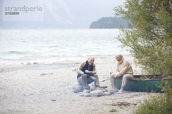 Germany  Bavaria  Walchensee  Senior couple sitting at campfire  grilling fish