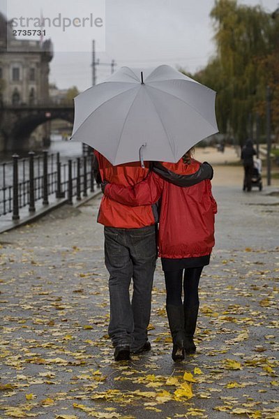 Rückansicht eines jungen Paares  das mit einem Regenschirm läuft.