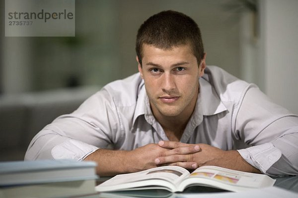 Ein junger Mann  der sich auf einen Schreibtisch mit einem offenen Buch stützt.