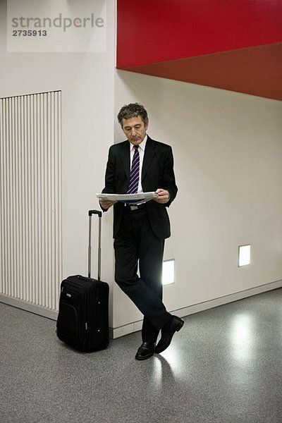 Ein Geschäftsmann  der die Zeitung liest und mit seinem Koffer wartet.