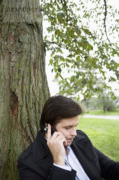 Ein Mann  der unter einem Baum sitzt und telefoniert.