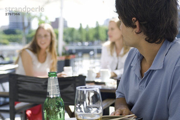 Junger Mann sitzt draußen im Café und schaut über die Schulter zu den jungen Frauen  die hinter ihm sitzen.