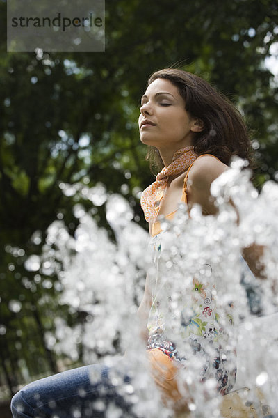 Junge Frau sitzend am Springbrunnen mit geschlossenen Augen  tiefer Blickwinkel