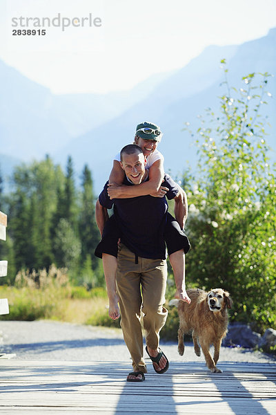 Ein Spaß-liebenden paar führt über eine Fußgängerbrücke in den Bergen mit dem Mann Huckepack der Frau und der Hund der Familie im Schlepptau  Kanada.